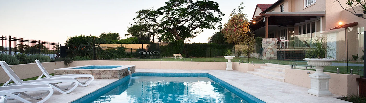 Backyard pool with lounge chairs, hot tub, and a large house surrounded by greenery.
