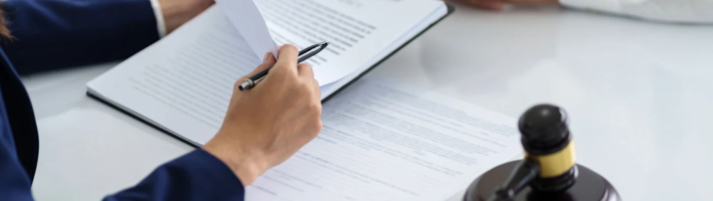 Two business people are sitting at a table, one holding paperwork with a gavel, and one with their hands folded in front of them.