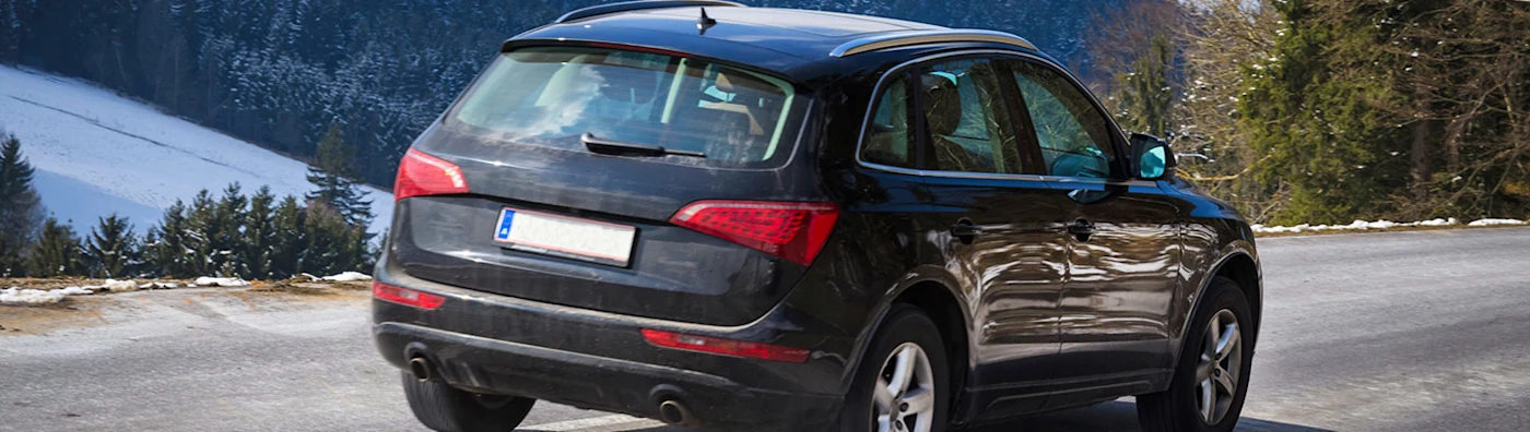 A black SUV drives along a mountain road with snow-covered hills and trees in the background.