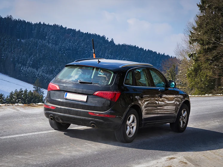 A black SUV drives along a mountain road with snow-covered hills and trees in the background.