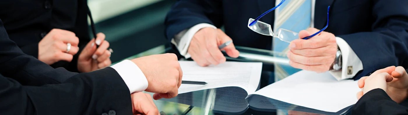 Close up image of business professionals in suits reviewing and discussing documents together at a glass conference table.