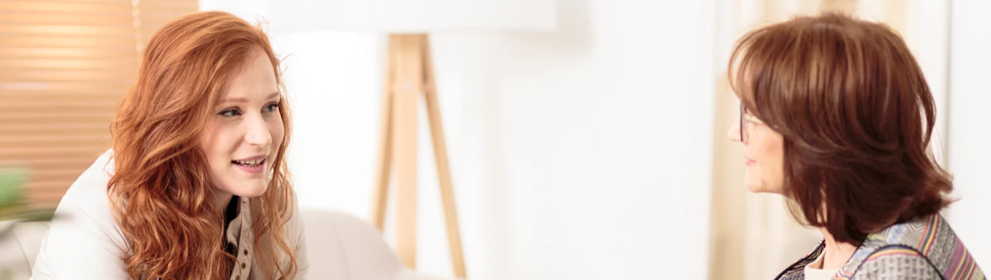 A woman sits on a couch in a mental health organization, talking with a therapist in a chair who holds a tablet computer.
