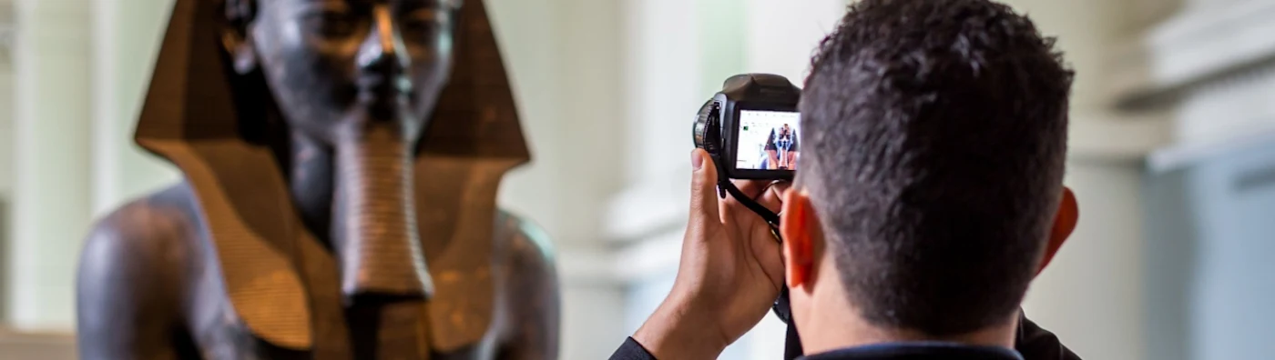 Tourist using conventional camera takes photo of Egyptian statue in a museum as he wonders about museum exhibition insurance.