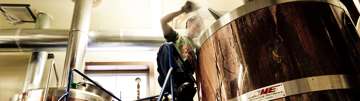 Worker brewing in a facility, surrounded by large stainless steel and wood-paneled equipment.