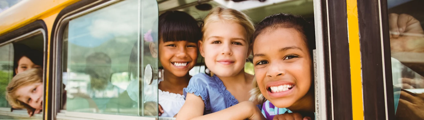 Smiling children look out the window of a yellow school bus on a sunny day.