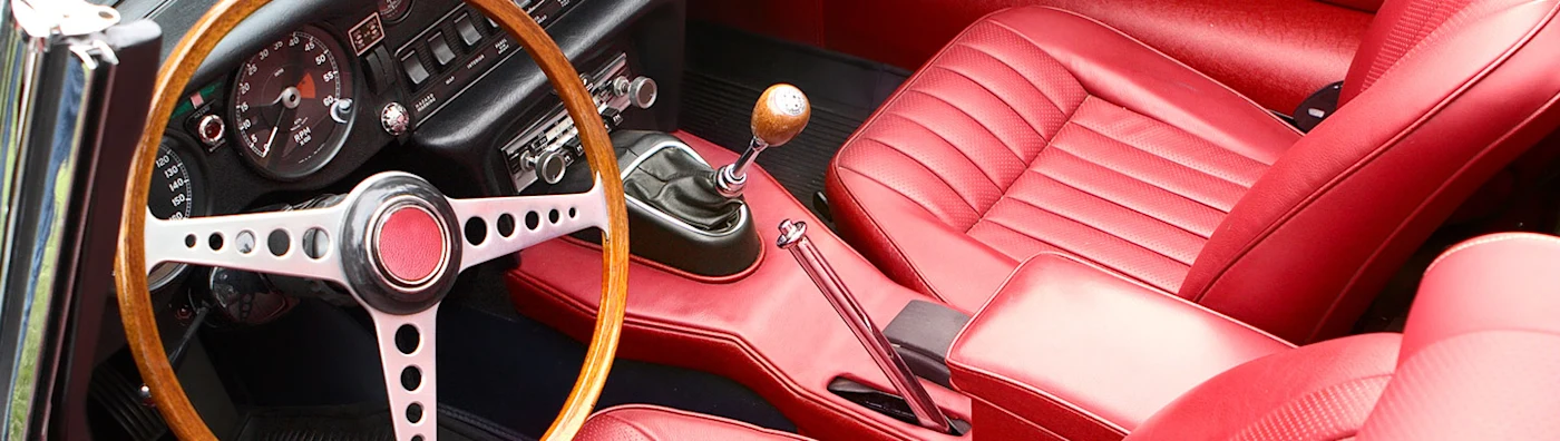 Close up image of the interior of an antique vehicle with red leather seats, a wood steering wheel, and manual gear shift.
