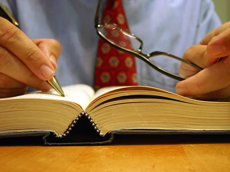 A close-up of a person in business attire holding a pen and glasses, poised over an open book for study or annotation.