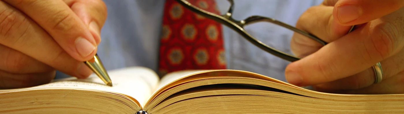 A close-up of a person in business attire holding a pen and glasses, poised over an open book for study or annotation.