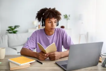 a teenage boy wearing headphones in a purple tshirt reading a book whilst looking at an open laptop. 