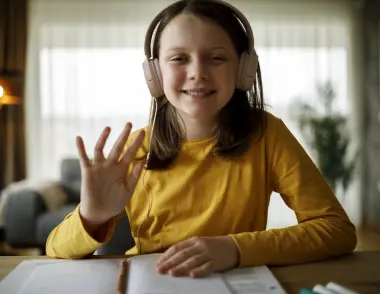 Young girl in a yellow top sitting at a computer with headphones waving to her online tutor