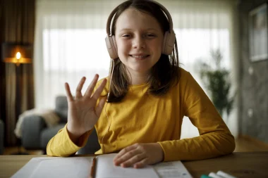 Young girl in a yellow top sitting at a computer with headphones waving to her online tutor.