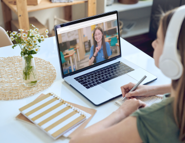 A teenage girl, wearing headphones, sits at a dining table with an open laptop in front of her.  On the screen of the laptop an Explore tutor is smiling back at her.