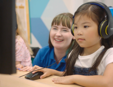 A young girl sits at a computer working with their tutor behind them assisting.