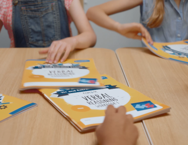 A series of 11 Plus workbooks on a table with children reaching out for them.