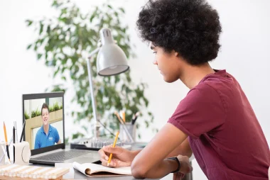 A young black teenager sits at a desk with an open laptop and notebook in front of him.  On the screen there is an Explore tutor smiling back at him.