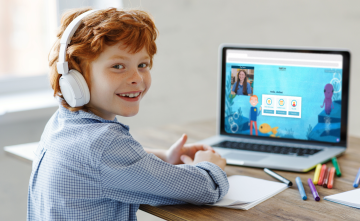 A child is smiling at the camera while sat in front of a laptop with a tutor and learning tool on screen.