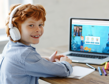 A child is smiling at the camera while sat in front of a laptop with a tutor and learning tool on screen.