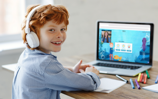 A child is smiling at the camera while sat in front of a laptop with a tutor and learning tool on screen.