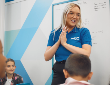 A young female tutor stands in front of a white board as she presents to a class of children.