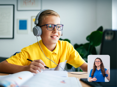 A cheerful young student with headphones participates in an online class, engaging with a tutor visible on the laptop screen, while sitting at a well-organised study area.