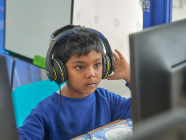 A young boy sits looking at a computer with headphones.