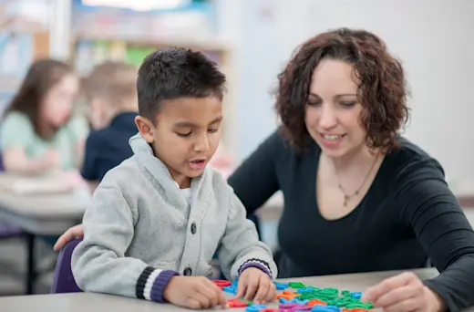 A teacher is helping a student with his toys at his table.