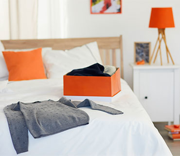 A gray pullover and an orange box full of clean clothes on a bed with a white duvet