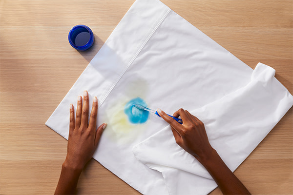 A person using a toothbrush to pretreat a stain with Tide liquid detergent on a white shirt