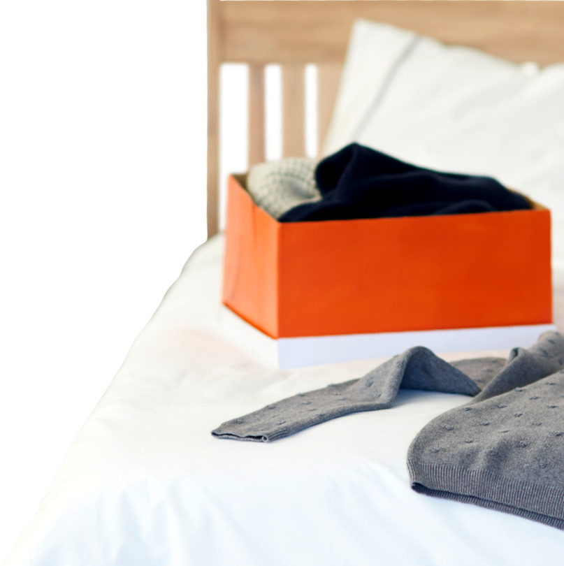 A gray sweater and an orange box full of clean clothes on a white bed
