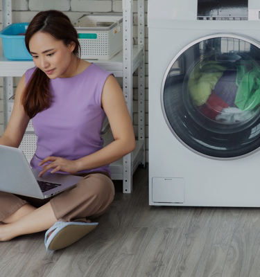 A young woman sitting on the floor with a laptop on her legs next to a washing machine full of clothes