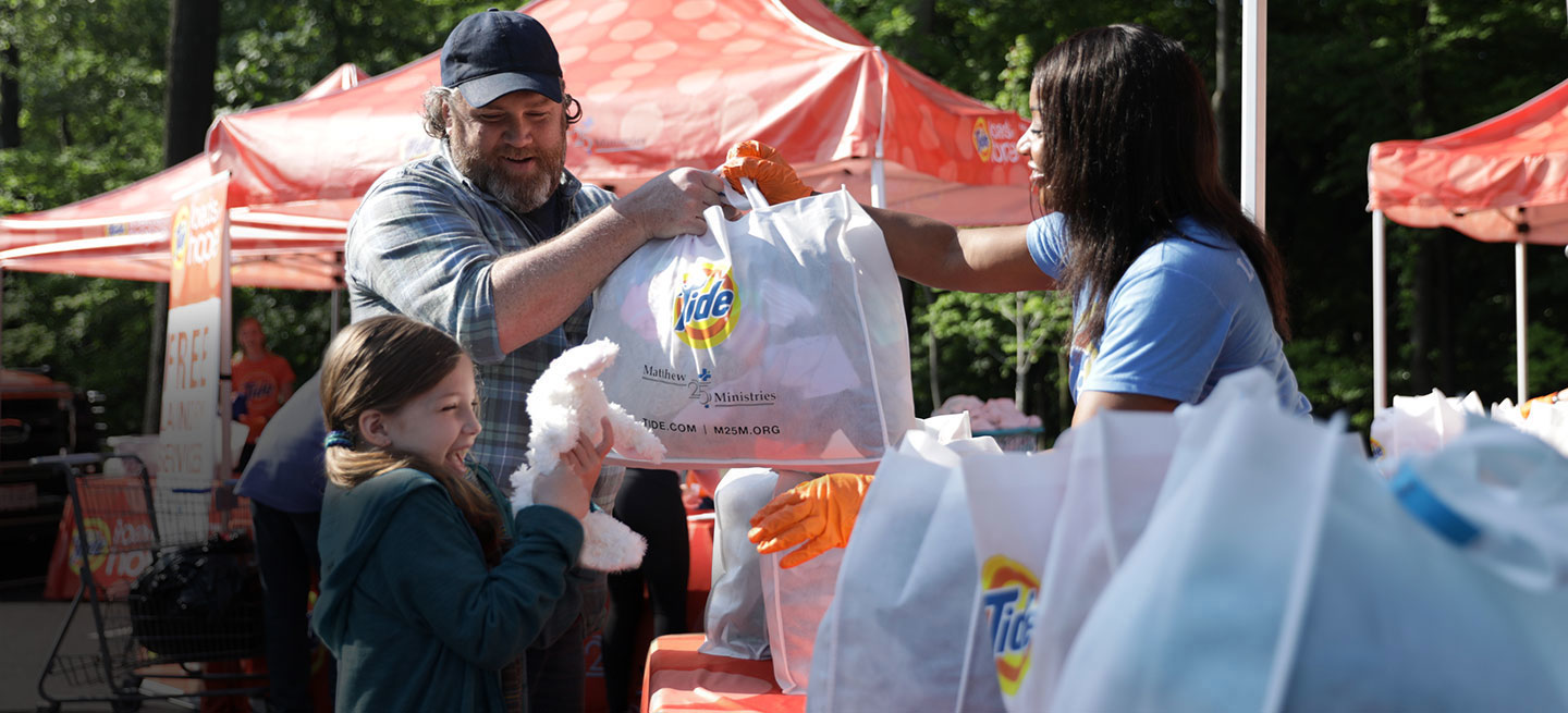 A man and a young girl handing over laundry to a woman working at a Tide tent set up after a natural disaster