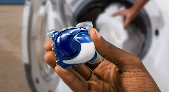 A hand holding a Tide PODS® capsule, while in the background, another hand is loading a washing machine