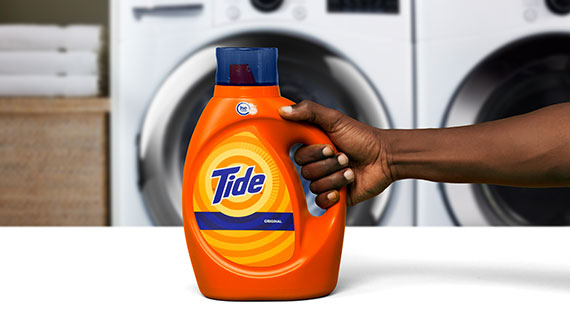 A hand holding a bottle of Tide Original liquid laundry detergent in front of a white washing machine