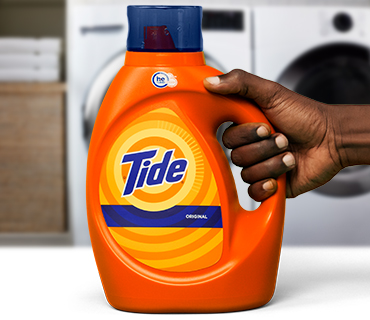 A hand holding a bottle of orange Tide Original liquid laundry detergent in front of a white washing machine