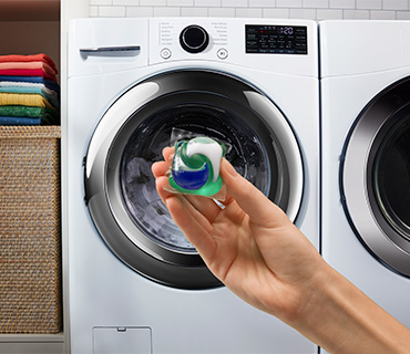 A hand holding a Tide PODS capsule in front of a white washing machine