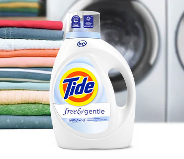 A white bottle of Tide Free and Gentle liquid laundry detergent in front of colorful folded clothes and a washing machine