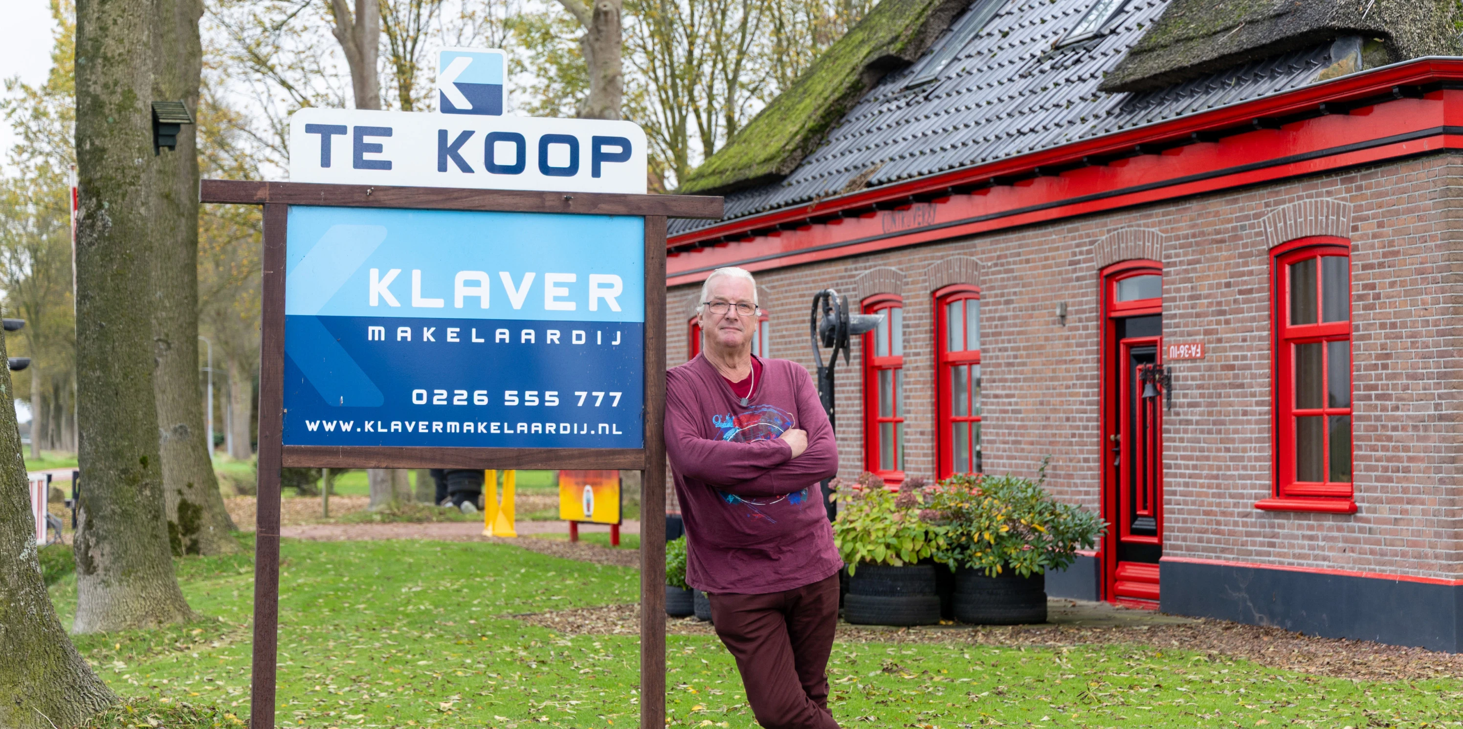 Verkoper Frank Appel staat voor zijn bijzondere woonboerderij geleund tegen het tekoopbord van Klaver Makelaardij. 