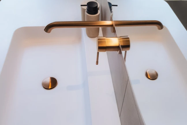 Detailbeeld van ingebouwde messing kranen in witte muur boven badmeubel van hout met witte, ingebouwde wastafel.