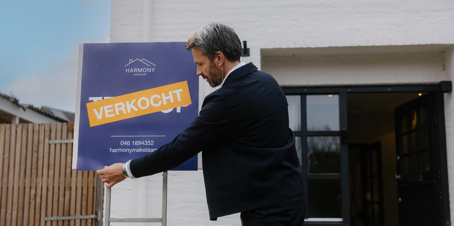 Een mannelijke makelaar plaats een verkochtbord in de voortuin van een witte moderne woning.