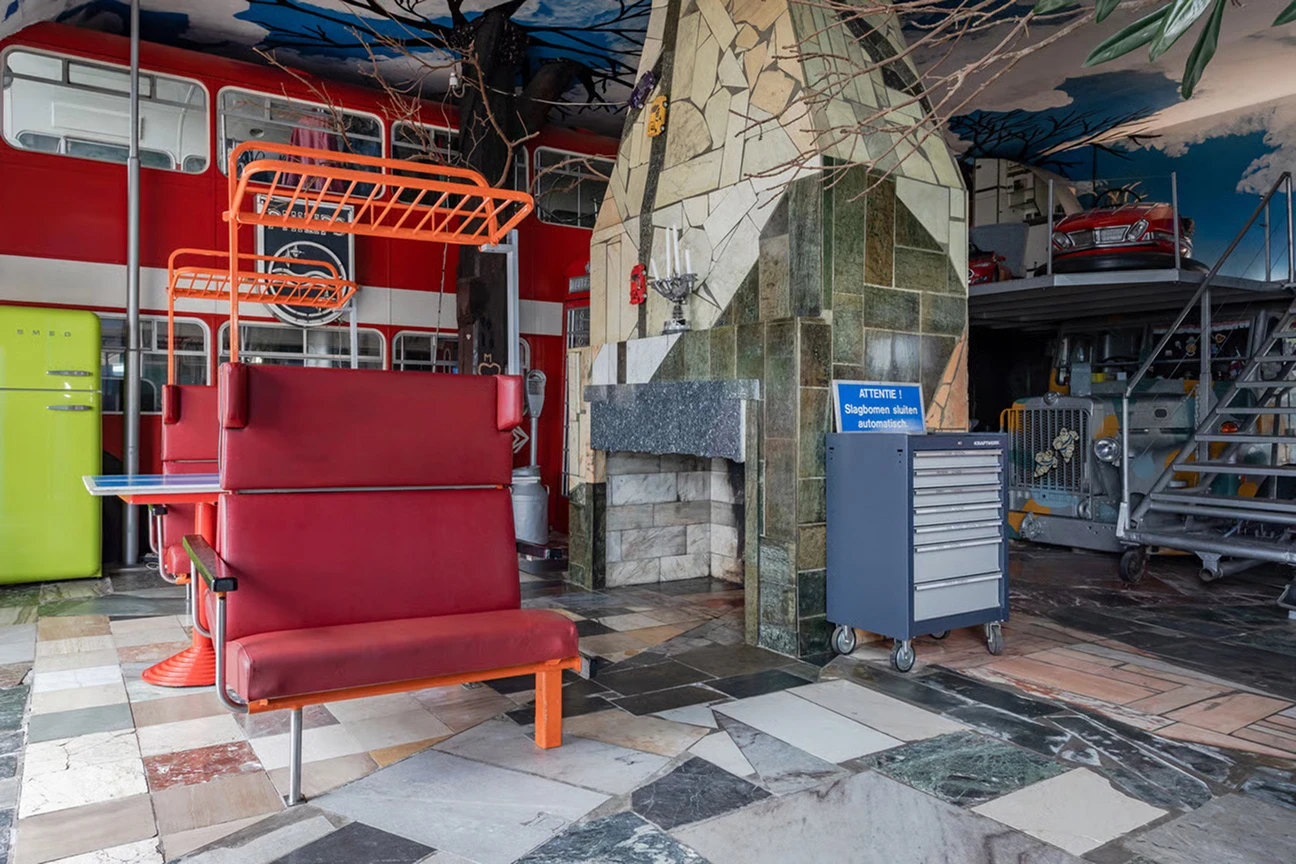 In de woonkamer staan rode banken uit een treinstel voor een grote, stenen houtkachel. Aan de achterzijde van de houtkachel is de voorzijde van een oude vrachtwagen uit het leger te zien.