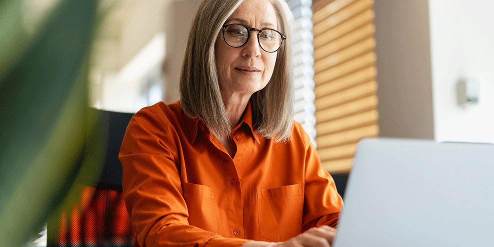 Vrouwelijke makelaar met halflang grijs haar en bril  werkt op laptop achter bureau.