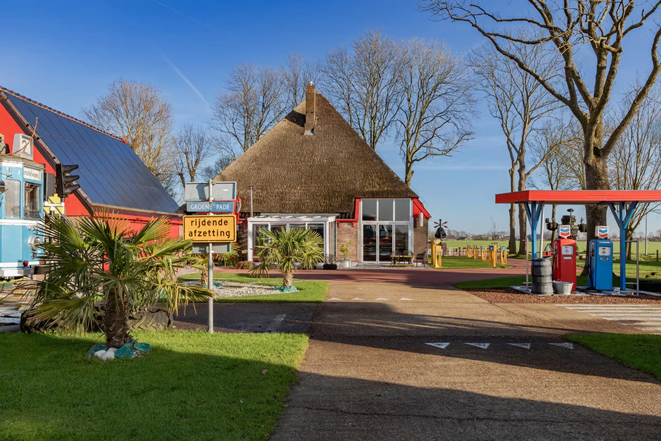 Rechts op het woonerf voor de stolpboerderij staat een oud tankstation met daarvoor een zebrapad. Links op de voorgrond is nog een deel van een lichtblauwe tram te zien. 