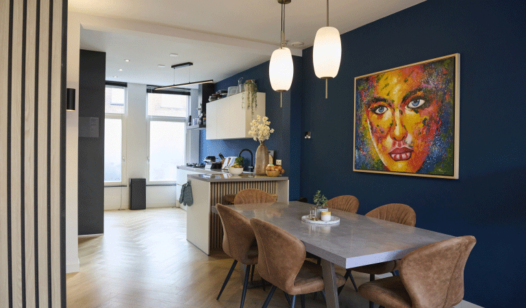 Een blik op de eettafel met op de achtergrond de keuken. Aan de donkerblauwe muur hangt een kleurrijk schilderij.