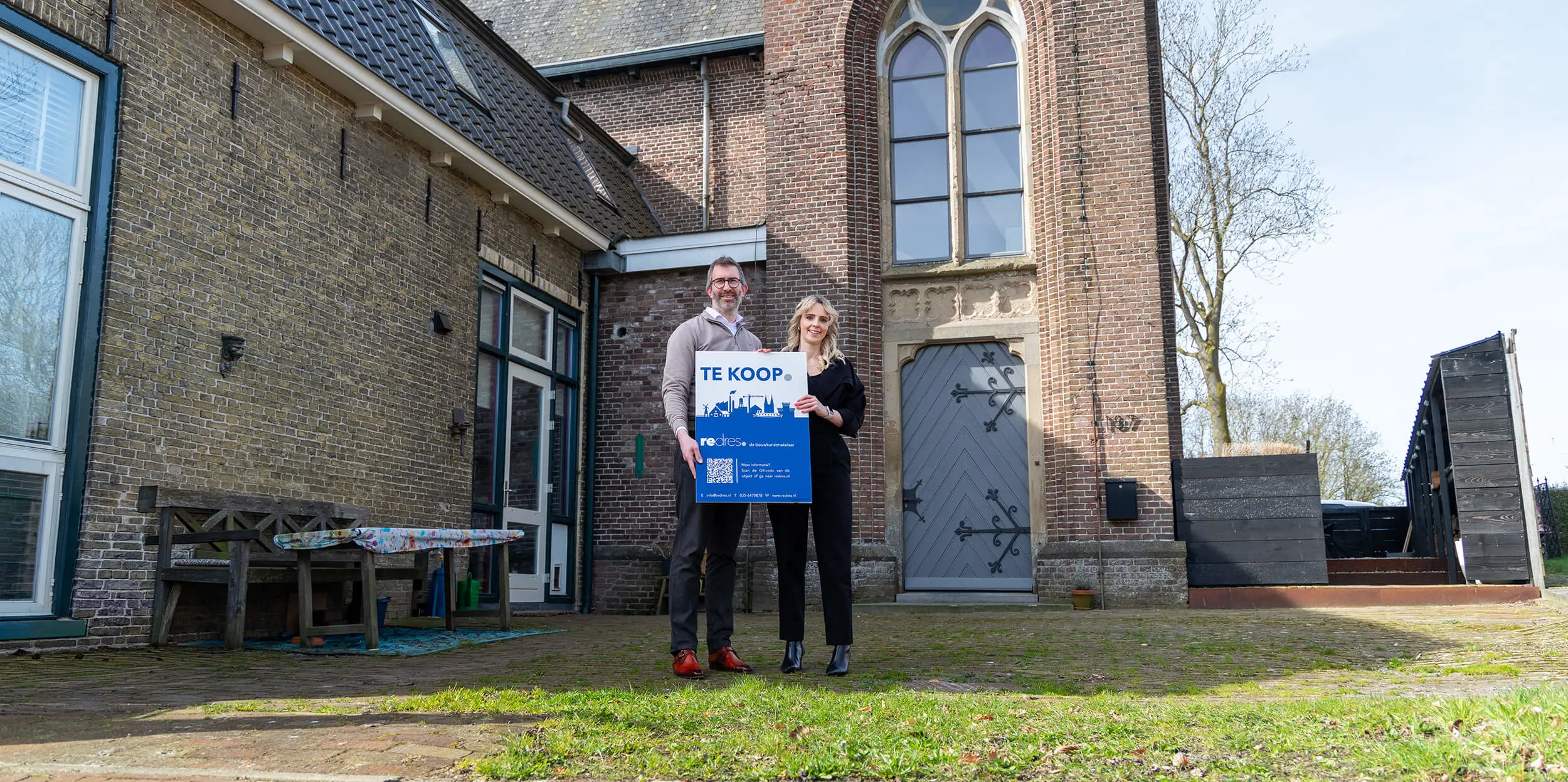 Katy en Rolf staan met een Tekoopbord voor hun kerk in Kuinre, die ze verbouwden tot woning. 