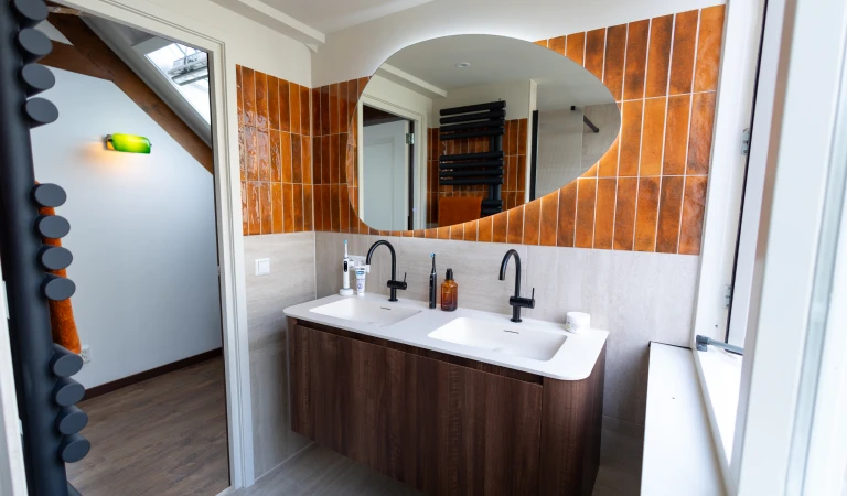 Doorkijkje vanuit de badkamer naar de overloop, met links op de voorgrond de designradiator en rechts het dubbele wastabelmeubel.