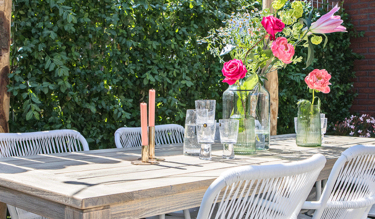 Buitenbeeld van vergrijsde houten eettafel met vier witte stoelen en een vaas bloemen op tafel. Op de achtergrond een groene haag.