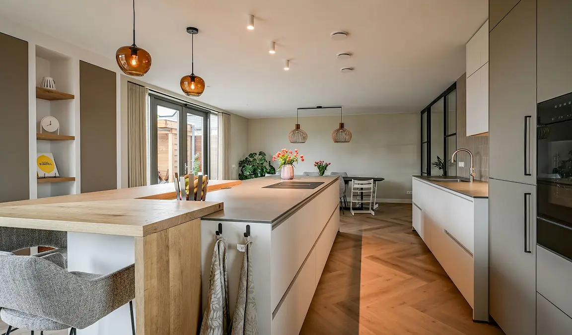 Ruime , witte-grijze keuken met grijs aanrechtblad en lichthouten zitbar aan kookeiland in woonhuis.