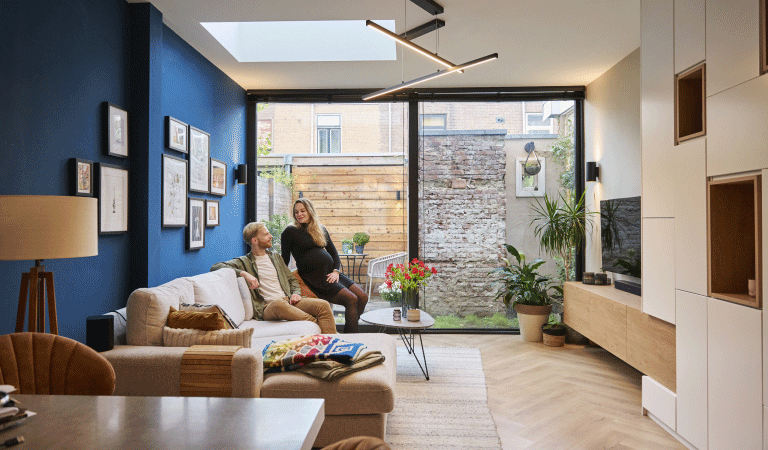 Djoeke en Goffe-Jan zittend op de bank in hun woonkamer. Op de achtergrond is de tuin te zien via de glazen schuifpui. De linkermuur is donkerblauw.