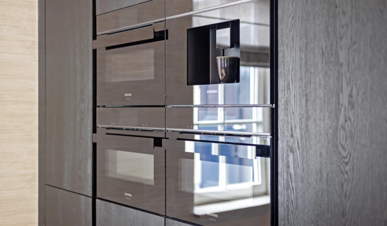 Voorbeeld drie ovens en ingebouwde koffieautomaat in keukenwand met donkerkleurige fronten.
