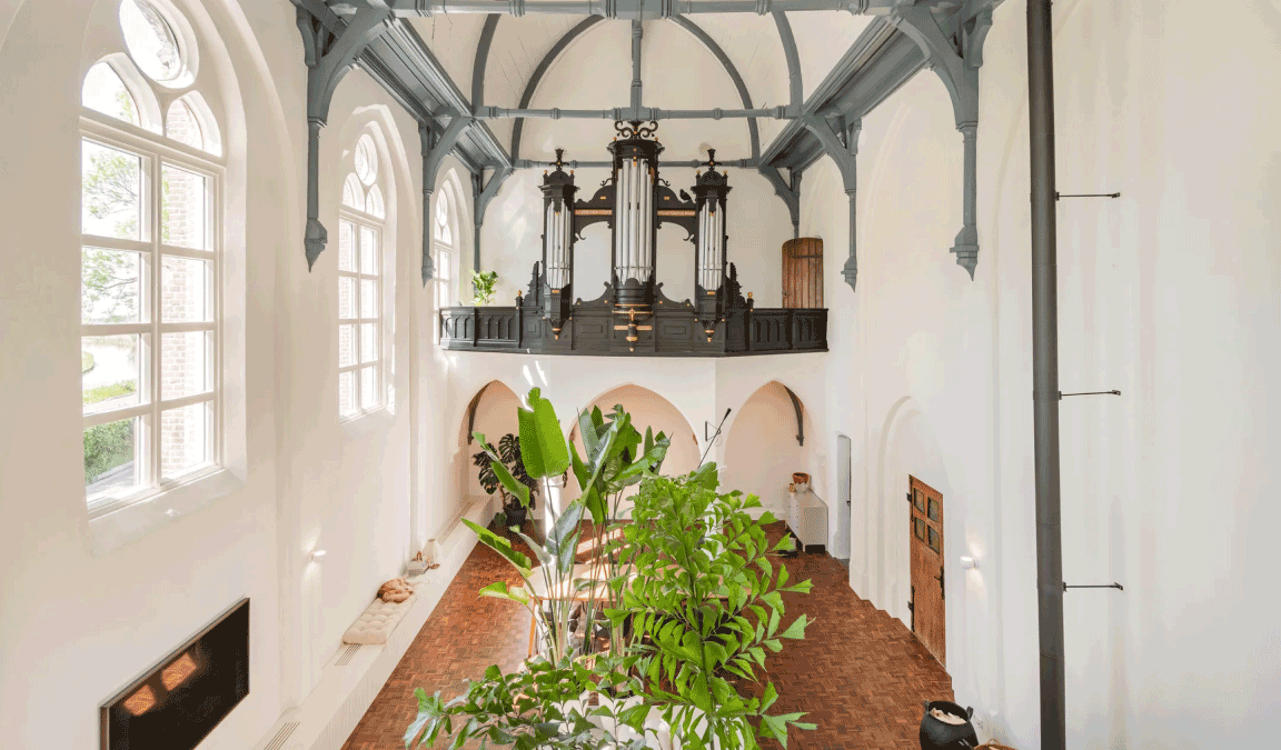 Een blik op de binnenkant van de kerk, het gedeelte dat verbouwd is tot woonkamer met witte muren. Het hoge plafond en het kerkorgel zijn behouden. 